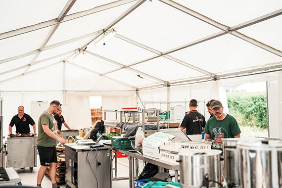 Six male Abbotts event staff arranging equipment within a long marquee tent, performing a range of tasks and in conversation with one another.